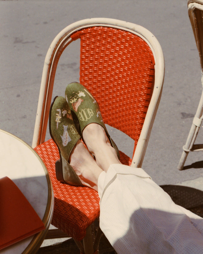 Wearing white pants, a person relaxes outdoors with feet in green Needlepoint House Shoes on a red woven chair beside a round table.