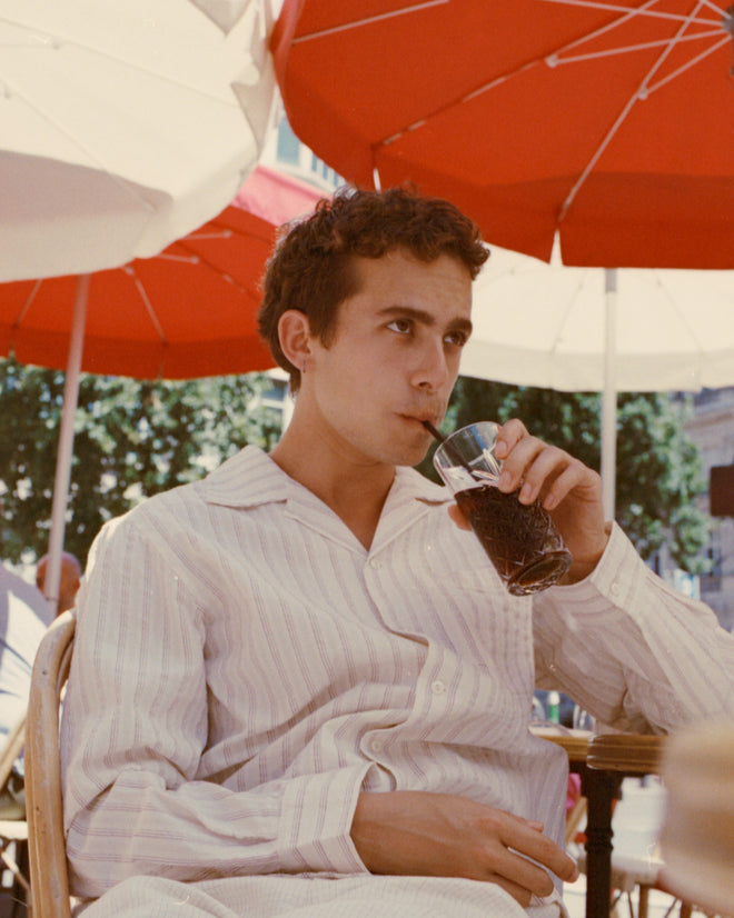 A young man wearing a Lavender Sail Shirt and striped pajamas sips soda through a straw at an outdoor café beneath red umbrellas.