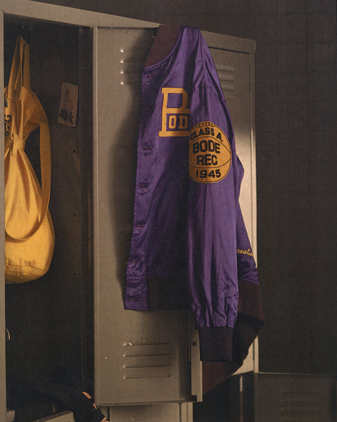 The Athlete's Rally Jacket, a vintage purple basketball championship jacket, hangs on a gray locker next to a yellow bag.