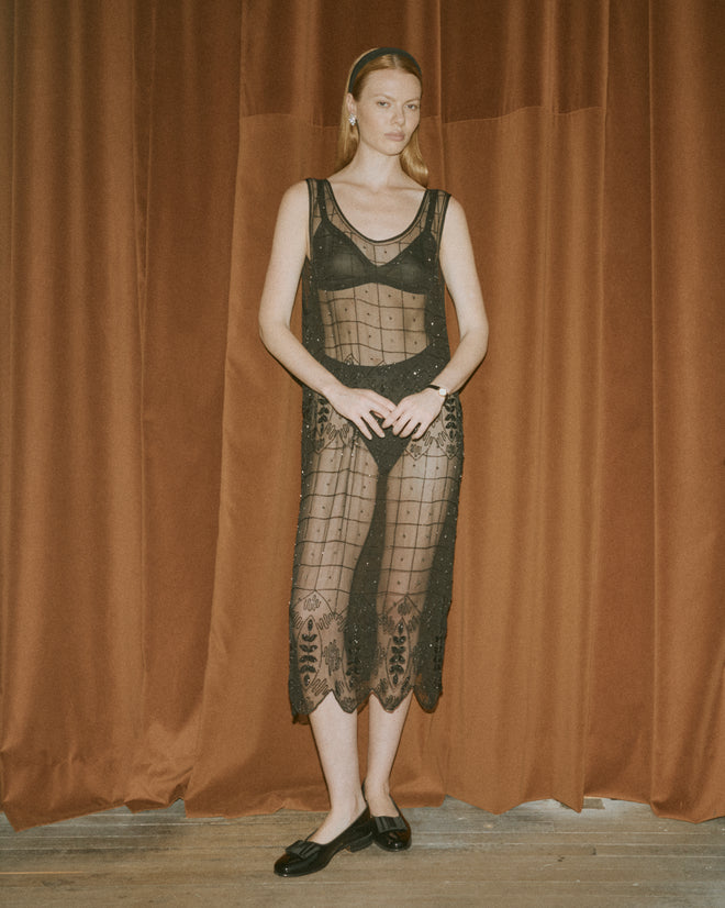 A woman wearing the Beaded Grid Vine Dress in black, a sheer silk crepe piece with delicate geometric vine embroidery, stands before reddish-brown curtains.