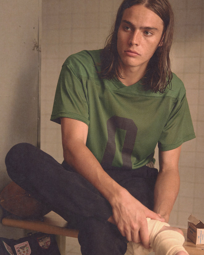 A young person with long brown hair wears a green Scrimmage Jersey, sitting indoors and gazing thoughtfully to the side.