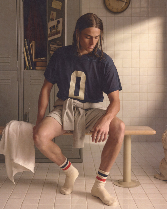 A young man wearing the Scrimmage Jersey - Navy and athletic gear sits on a locker room bench, looking down, with a towel draped beside him.
