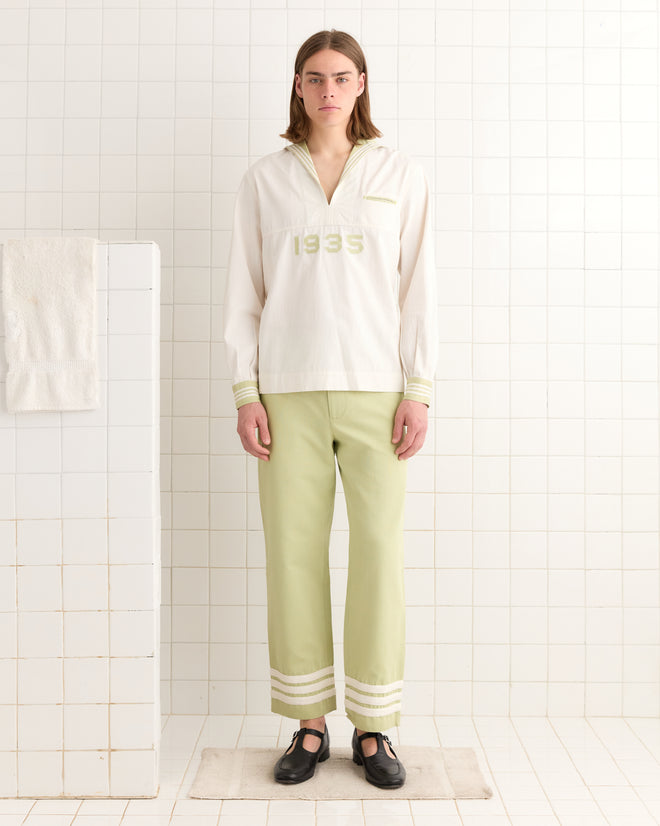 A person in a 1935 Cricket Shirt with an antique white finish and subtle sailor collar, paired with light green pants and black shoes, stands in a tiled room.