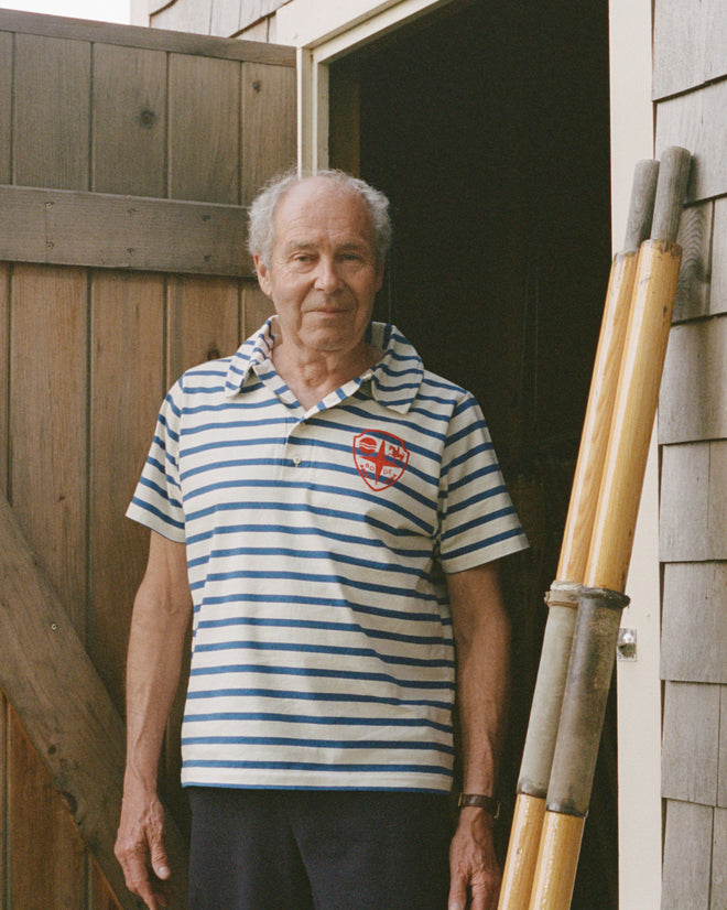 An older man wearing the Margate Polo in French Navy stripes stands by a wooden fence and shed, holding a tall wooden ladder.
