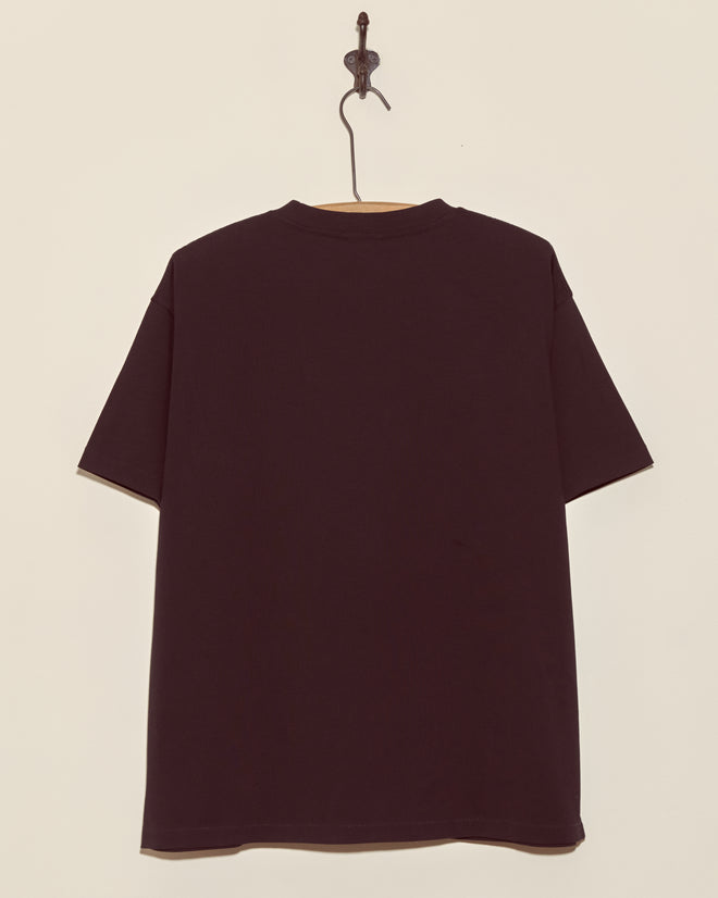 The Skeleton Tee, inspired by vintage skeleton designs, is a plain black T-shirt displayed from the back on a hook against a beige wall.