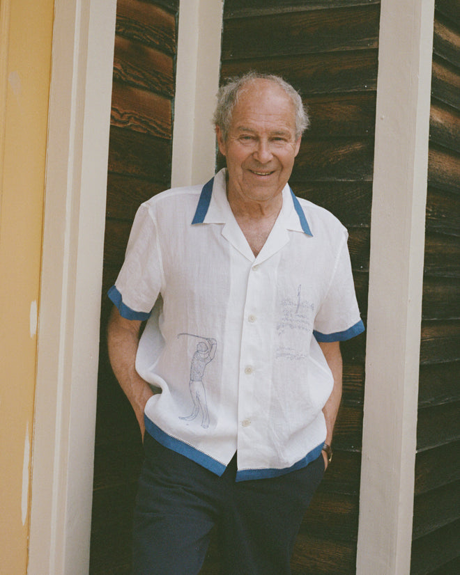 An older man with gray hair, smiling and standing against a wooden wall, wears the Club Champion Short Sleeve Shirt—a crisp white golf shirt with blue trim.