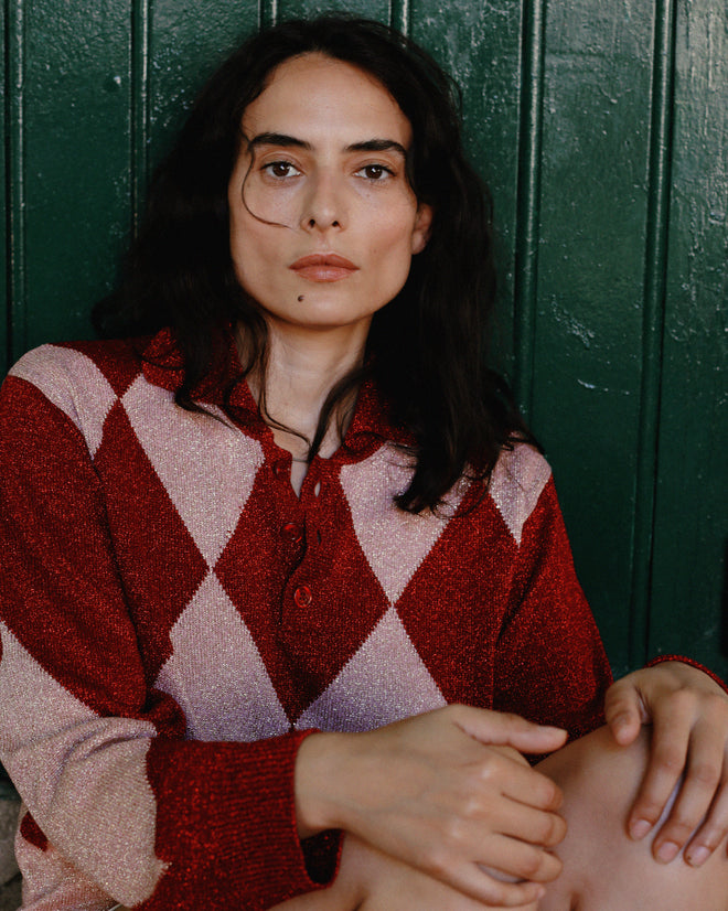 A person with long dark hair wearing the Jockey Polo in a red and pink argyle pattern sits in front of a green paneled wall.