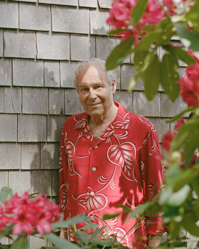 An older man wears the Creeping Begonia Long Sleeve Shirt, with a midcentury botanical print, standing by a shingled wall and blooming pink flowers.