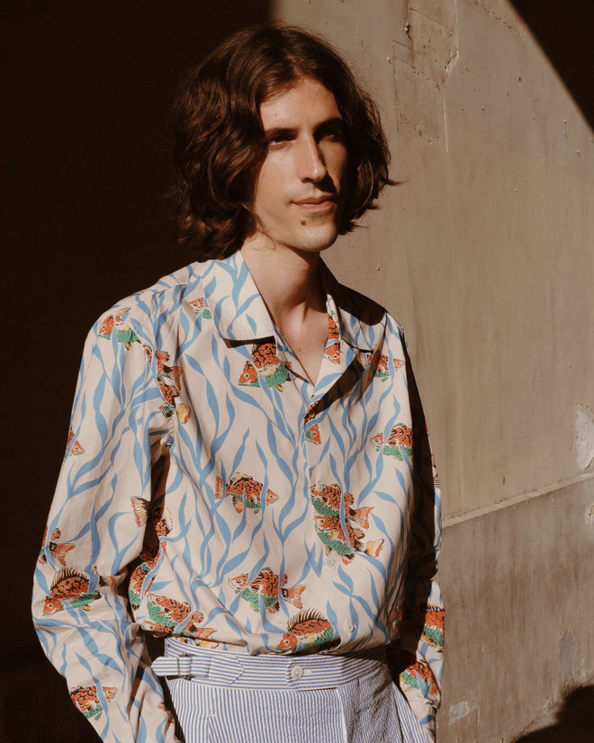 A person with wavy brown hair wears the Swimmers Long Sleeve Shirt, featuring a vibrant midcentury print, standing in sunlight against a textured beige wall.