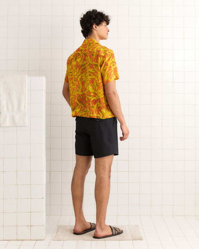 A man stands in a tiled room, facing away, wearing the Abstract Foliage Short Sleeve Shirt—1970s inspired with a yellow and red pattern made from 100% silk deadstock fabric—paired with black shorts and sandals.