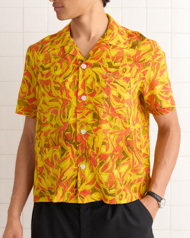 A person wearing the Abstract Foliage Short Sleeve Shirt, inspired by the 1970s and crafted from deadstock fabric, stands against a white tiled wall.