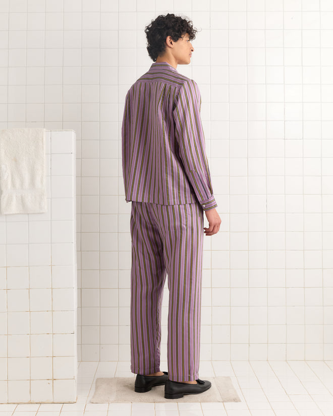 A person stands in a tiled room facing away, wearing the Alumni Stripe Long Sleeve Shirt - Purple, featuring bold purple, green, and black stripes with a retro vibe.
