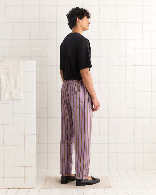 A person stands sideways in a tiled room, wearing a black shirt, Alumni Stripe Pajama Pants- Purple, and black shoes.