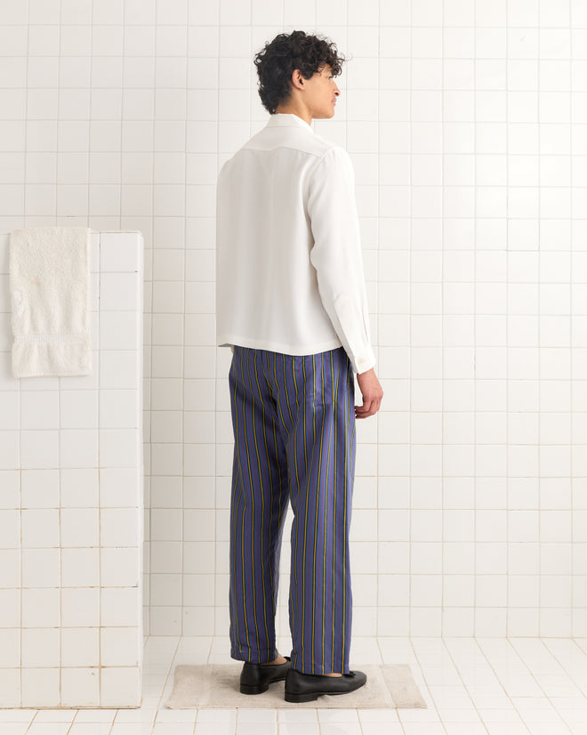 A person stands in a tiled room, facing away from the camera, wearing a white shirt and Alumni Stripe Pajama Pants- Blue.