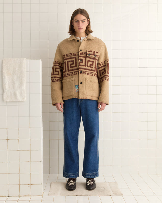 A person stands in a tiled room wearing the Apollo Ornament Jacket - M/L, a unique beige wool piece with a Greek meander motif, styled with blue jeans and black shoes.