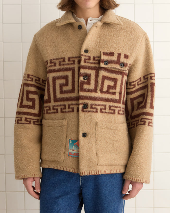 A person stands against a tiled wall wearing the Apollo Ornament Jacket - M/L, a unique wool piece with geometric brown patterns inspired by the Greek meander motif, paired with blue jeans.