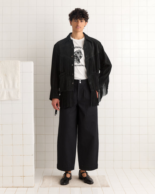 A person in black wide-leg pants, the Appalachian Jacket - Black with 1960s Western fringe details, and a printed white T-shirt stands against a tiled wall.