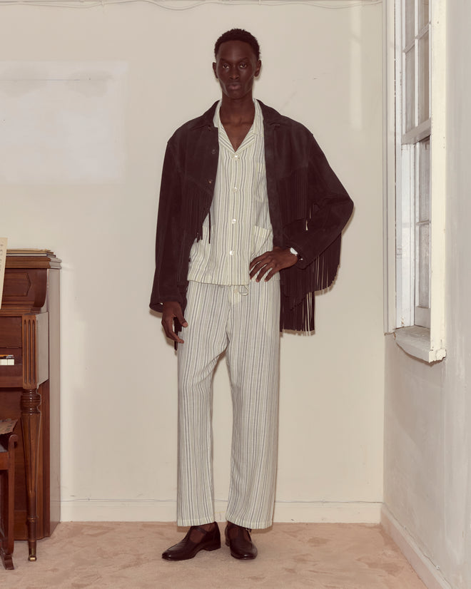 A person wearing the Appalachian Jacket - Black, featuring hand-cut fringe, stands indoors near a piano and a window.