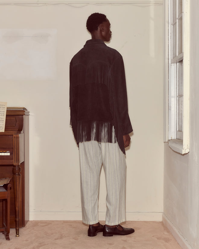 A person stands indoors, facing away, wearing the Appalachian Jacket - Black with hand-cut fringe and striped white pants.