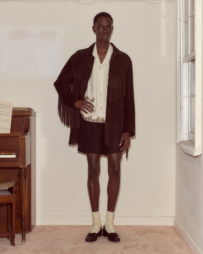 Indoors, a person wears the Appalachian Jacket - Brown (suede with hand-cut fringe), paired with a patterned shirt, black shorts, and loafers with socks.