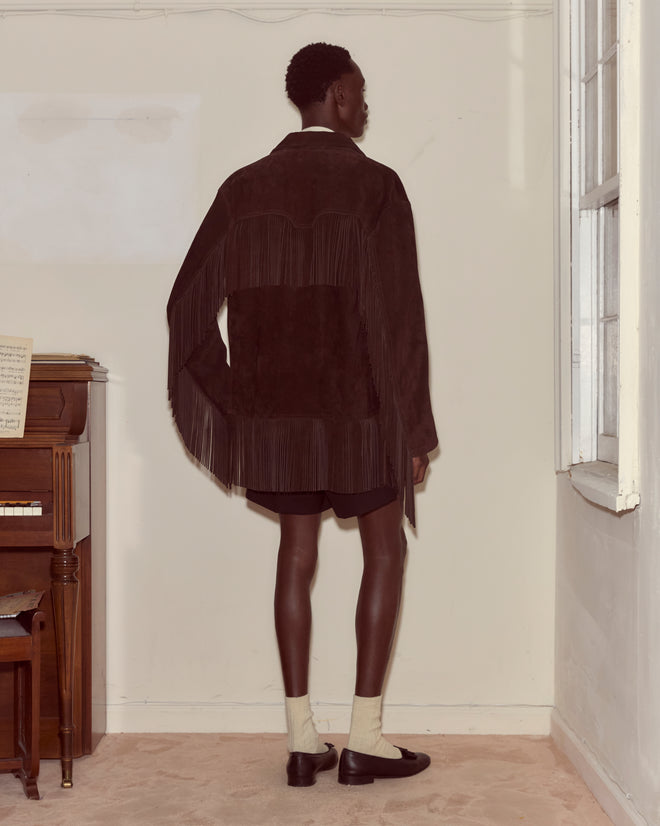 Indoors by a piano, person faces away wearing the Appalachian Jacket - Brown, a dark suede jacket with hand-cut fringe, paired with shorts, socks, and loafers.