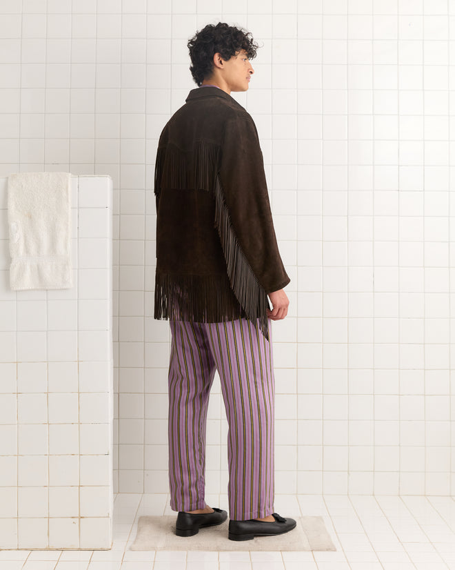 A person in the Appalachian Jacket - Brown with hand-cut fringe and striped pants stands sideways in a tiled bathroom.