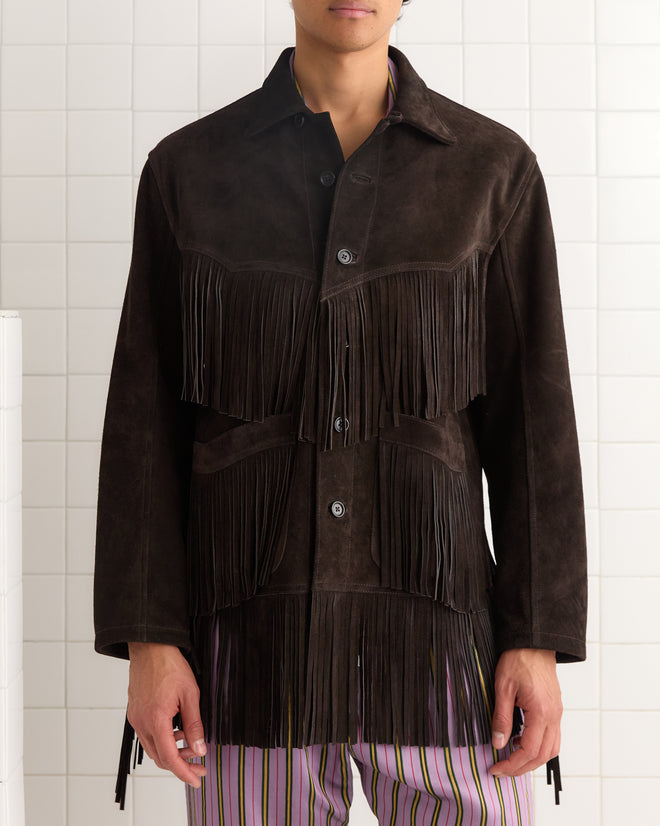 Person wearing the Appalachian Jacket - Brown, a dark suede piece with hand-cut fringe, paired with striped pants and standing against a white tiled wall.