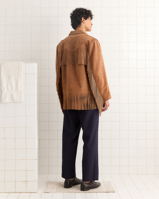 A person wearing the Appalachian Jacket - Light Brown with hand-cut fringe and dark pants stands in a white tiled room, facing away.