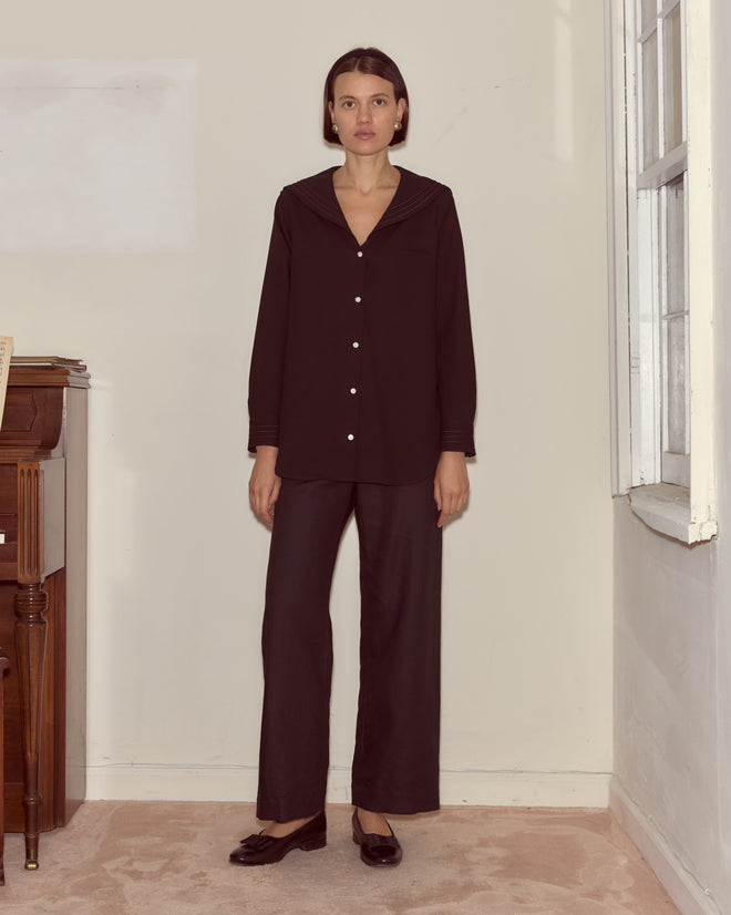 A woman stands indoors by a window, wearing the Astor Blouse in black wool crepe with a sailor collar, matching pants, and black shoes.