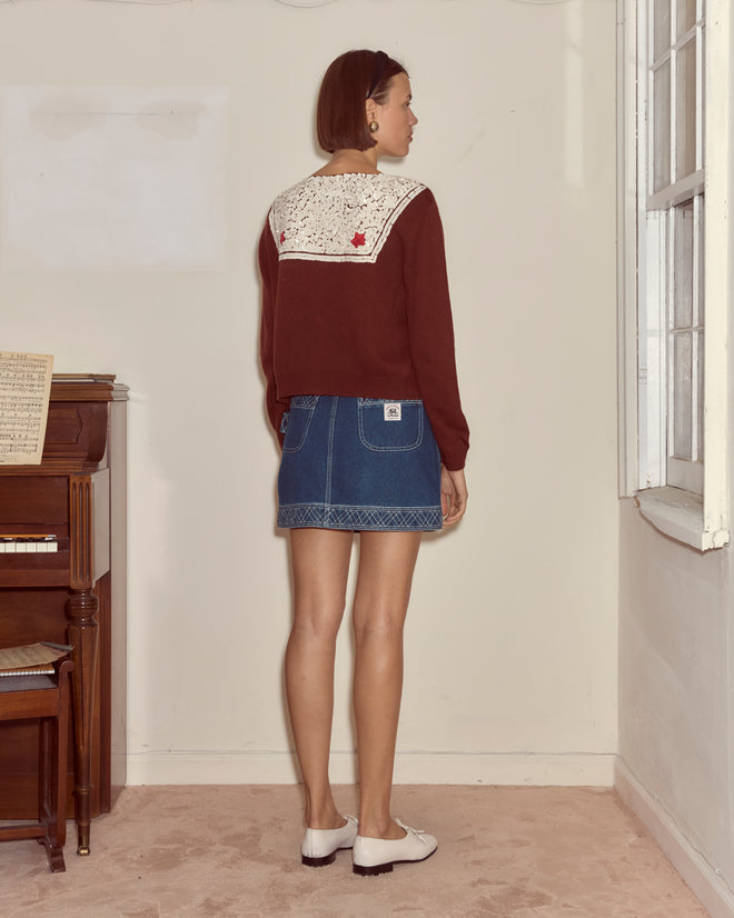 A woman stands indoors facing away, wearing the Astoria Cardigan in brown and a denim skirt, near a piano and window.