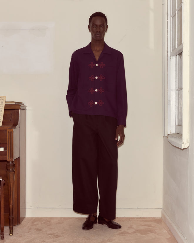 Someone stands indoors by a piano and window, wearing the Astor Trillium Shirt in purple with wide black pants.