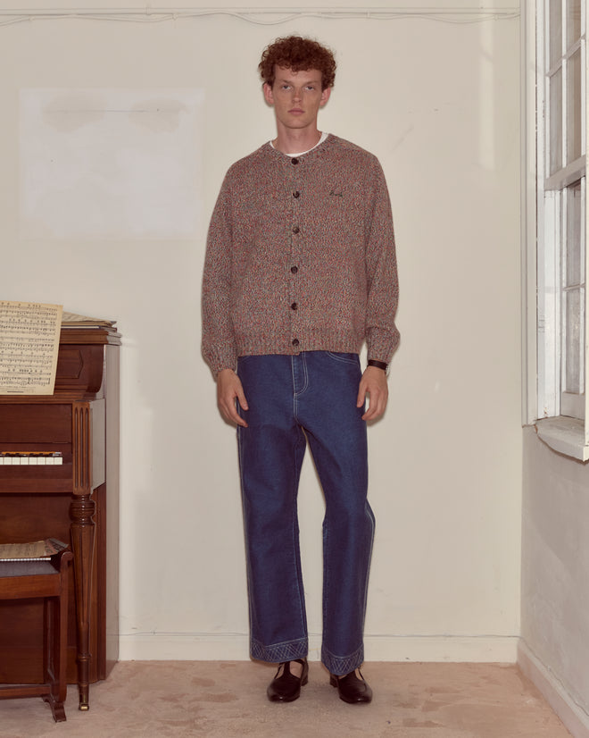 A person wearing the Atlas Cardigan and jeans stands indoors beside a piano and window, facing the camera.