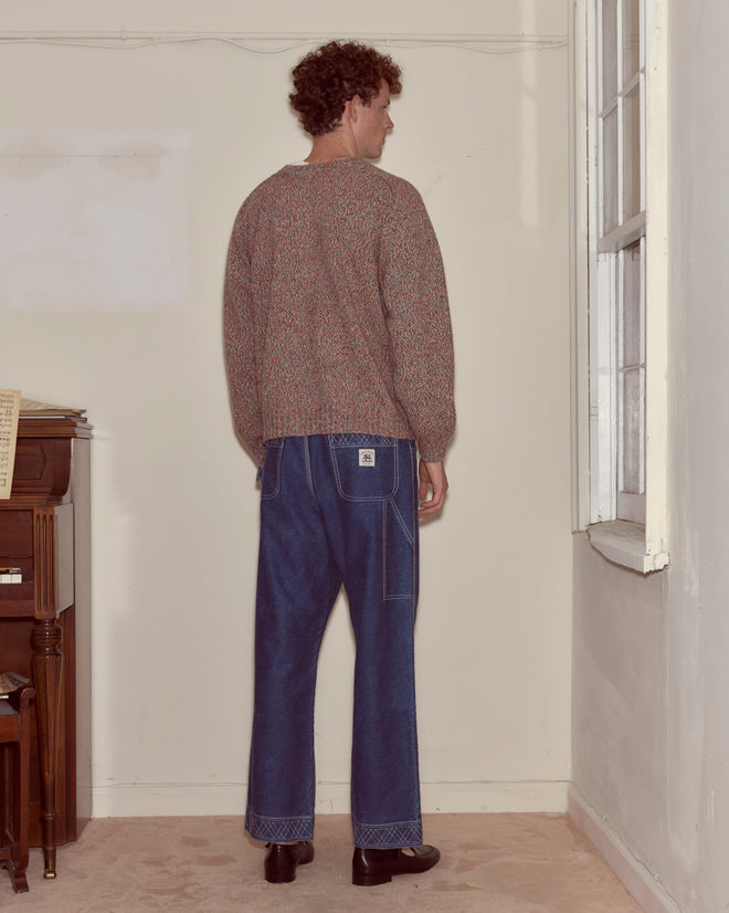 A man with curly hair stands facing a wall in a room, wearing blue jeans, black shoes, and the Atlas Cardigan.