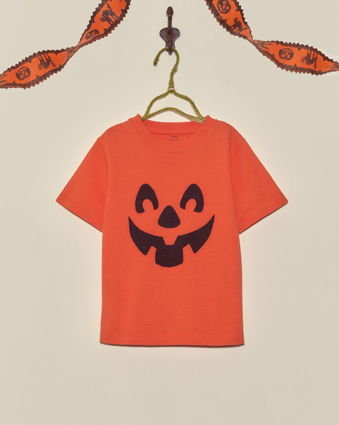 The Pumpkin Kids' Tee, featuring a bold black jack-o-lantern face, hangs on a hanger with Halloween garland above—making it the perfect festive vintage Halloween shirt for kids.