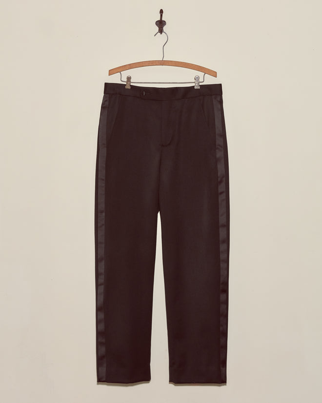 Barathea Tuxedo Trousers, crafted from 100% wool with a satin tuxedo stripe, hang on a wooden hanger against a plain beige wall.