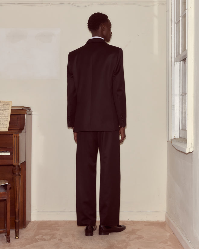 A man in a Barathea Peak Lapel Tuxedo Jacket stands facing a corner in a sparse room with a piano and window.