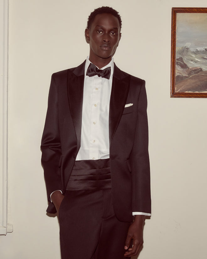 A man wearing the Barathea Peak Lapel Tuxedo Jacket and a bow tie stands indoors beside a framed painting on the wall.