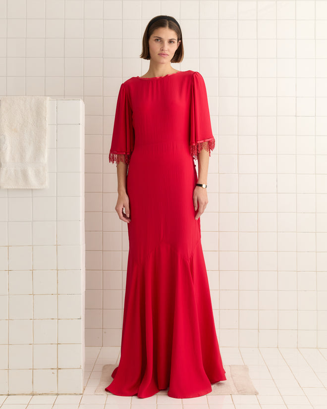 A woman in a Beaded Claret Gown - Red with fringed sleeves stands against a tiled bathroom wall.