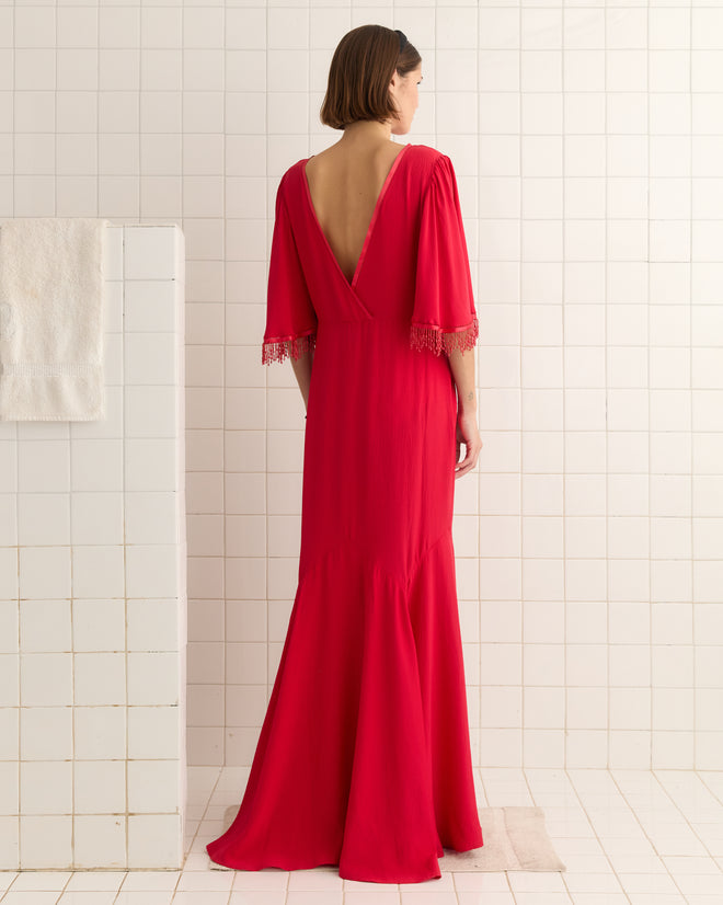 In a Beaded Claret Gown in red with a 1930s vibe and open back, a woman stands in a tile room beside a white towel hanging on a wall divider.
