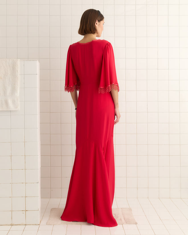 A person in a Beaded Claret Gown - Red, featuring fringed sleeves and a 1930s-inspired godet skirt, stands elegantly in a white tiled room.