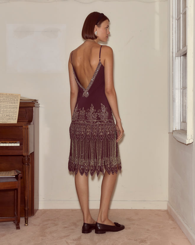 Woman in a vintage-inspired deep purple Beaded Epoque Dress with gold detailing stands by a piano, facing away in a softly lit room.
