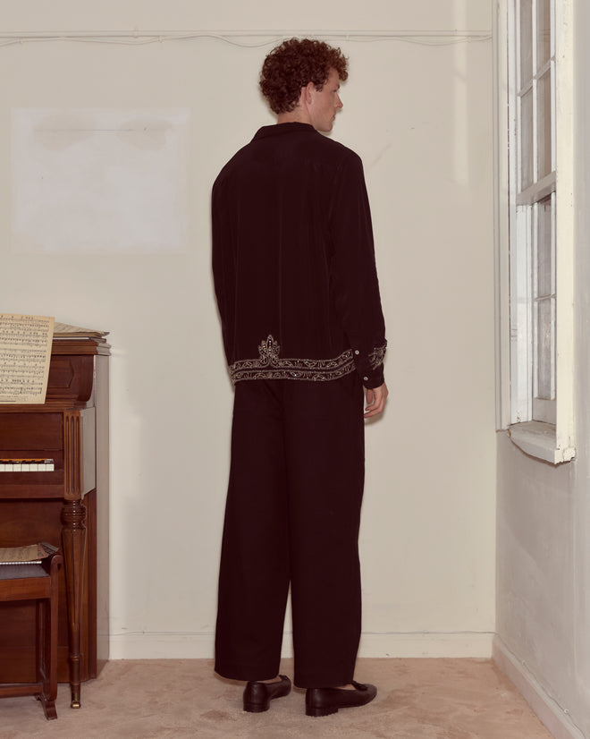 A man in a Beaded Epoque Long Sleeve Shirt faces a wall beside a piano with sheet music on it.