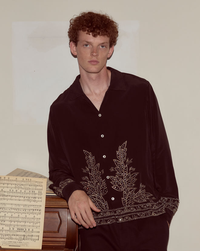 A young man wears the Beaded Epoque Long Sleeve Shirt as he leans on a piano with sheet music, set against a plain wall.