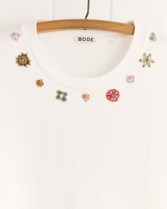 The Beaded Floral Necklace Tee hangs on a hanger, featuring colorful embroidered patches around the neckline. Crafted from 100% cotton, its unique textile details are inspired by vintage sari appliqués.