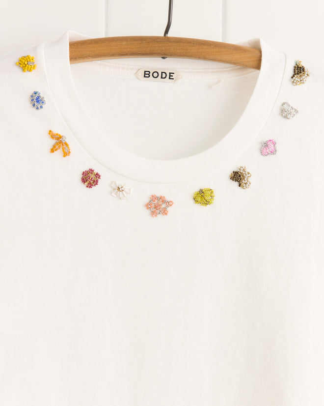 The Beaded Floral Necklace Tee, made from 100% cotton, features small, colorful embroidered accents around the neckline for a unique look and is displayed on a wooden hanger.
