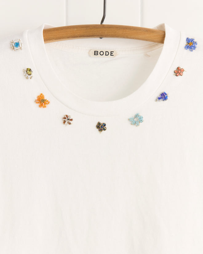 The Beaded Floral Necklace Tee by BODE is a white 100% cotton t-shirt with colorful beaded floral and gem embellishments inspired by vintage sari appliqués around the collar, shown on a wooden hanger.