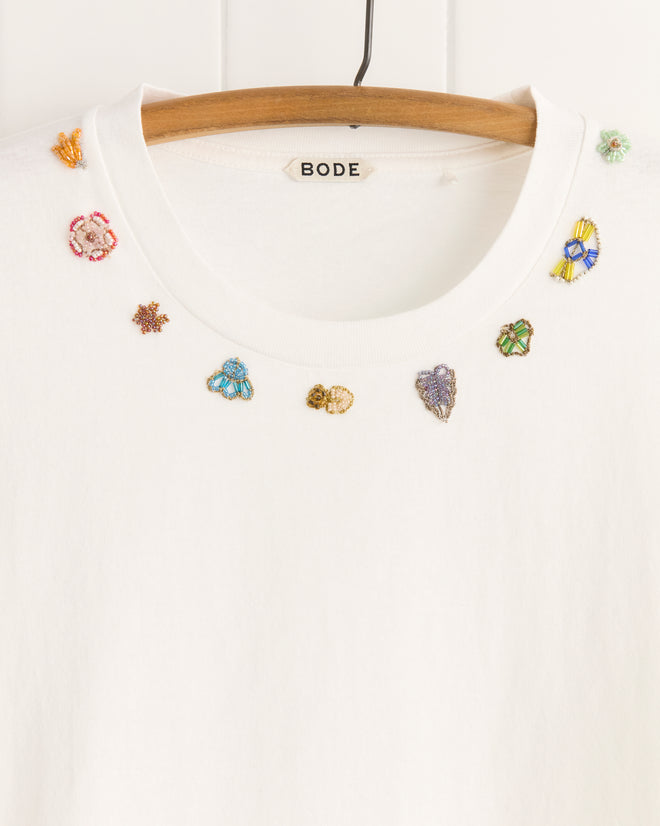 The Beaded Floral Necklace Tee by BODE is a white 100% cotton t-shirt featuring small, colorful beaded and embroidered motifs around the collar, with unique details inspired by vintage sari appliqués.