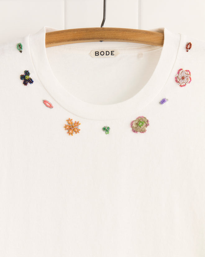 The Beaded Floral Necklace Tee, crafted from 100% cotton, features small colorful beadwork and vintage sari appliqués along the neckline, shown here hanging on a wooden hanger.