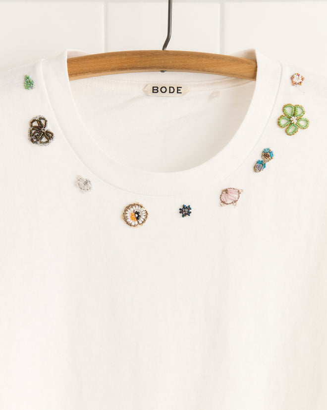 The Beaded Floral Necklace Tee is a white 100% cotton Bode t-shirt on a hanger, featuring colorful beaded embellishments near the neckline and unique textile details inspired by vintage sari appliqués.