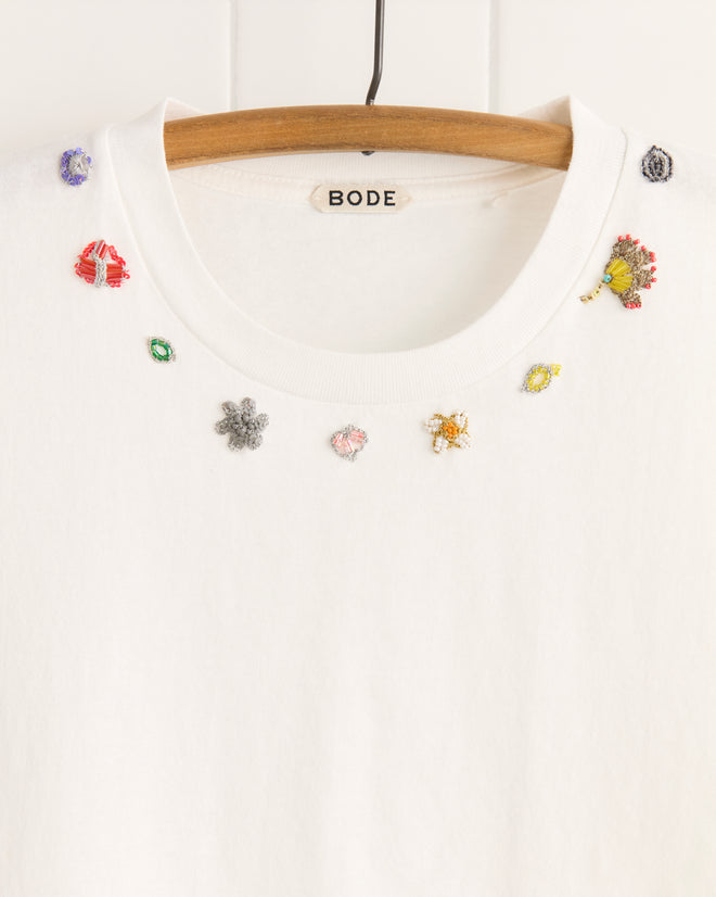 The Beaded Floral Necklace Tee is a white 100% cotton BODE shirt, adorned with small embroidered flowers and shapes at the neckline, inspired by vintage sari appliqués, displayed on a wooden hanger.